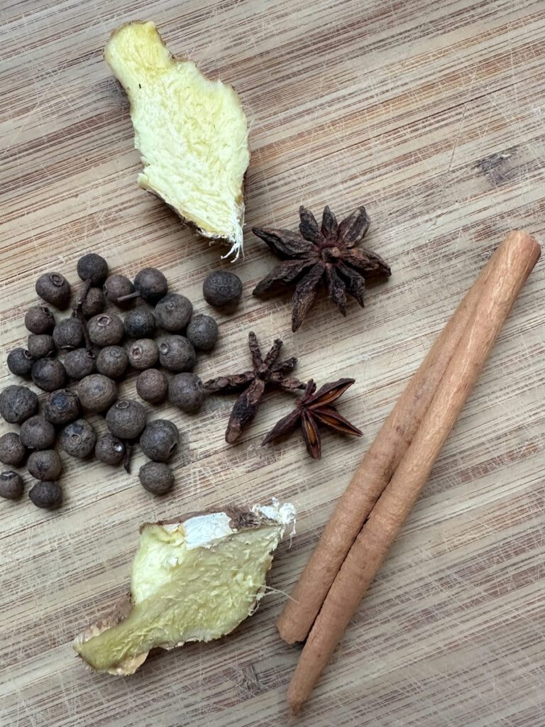 Warm spices including fresh ginger, all spice berries, star anise pods, and cinnamon stick on a wooden cutting board.