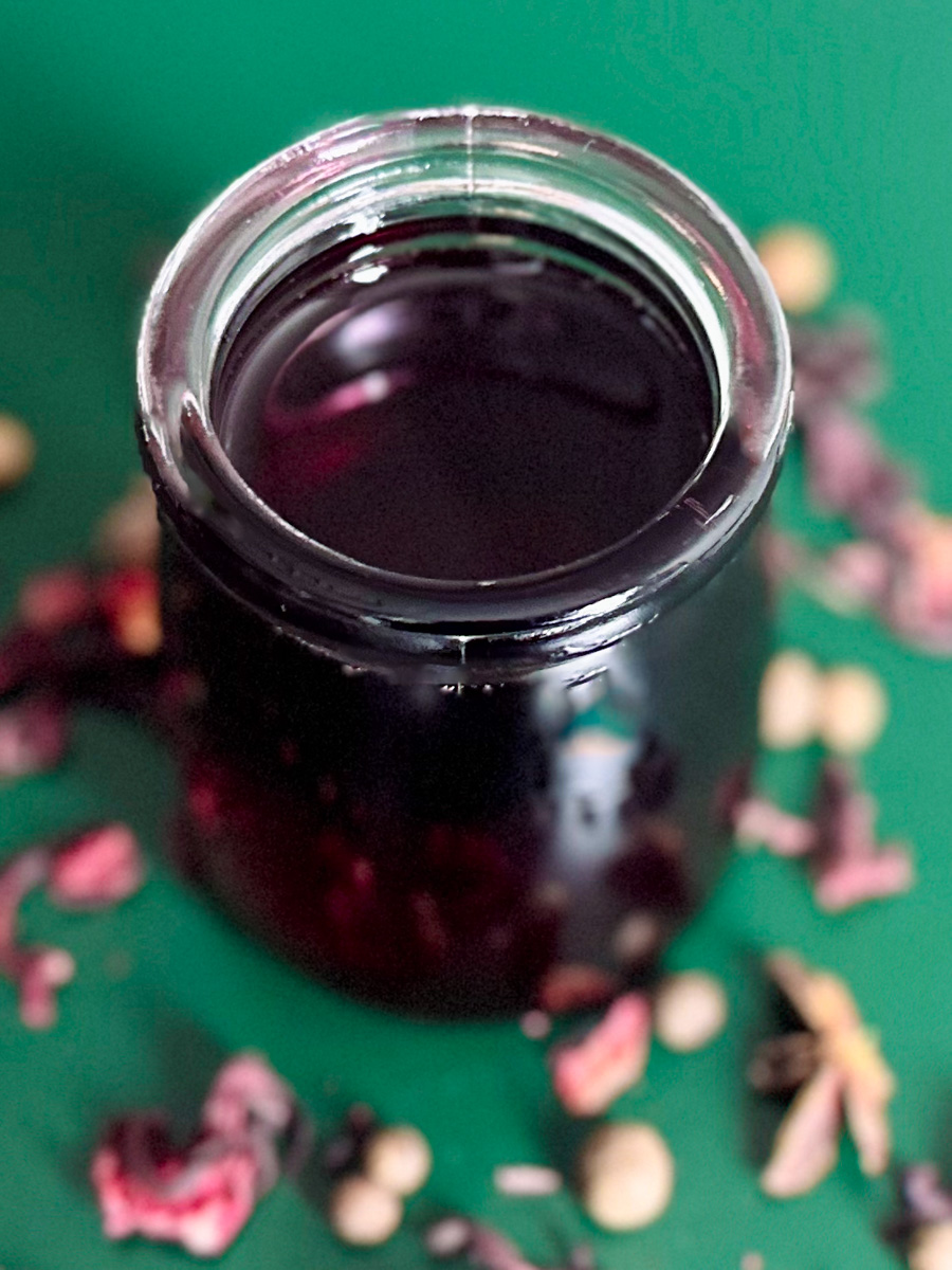 Sorrel syrup on a green background surrounded by dried hibiscus leaves and all spice berries.