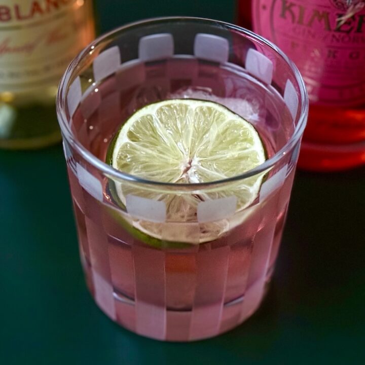 Pink Negroni Cocktail made with pink gin, lillet blanc, and lime setting on a green table with bottles of Lillet Blanc and Dry Vermouth bottles behind it.
